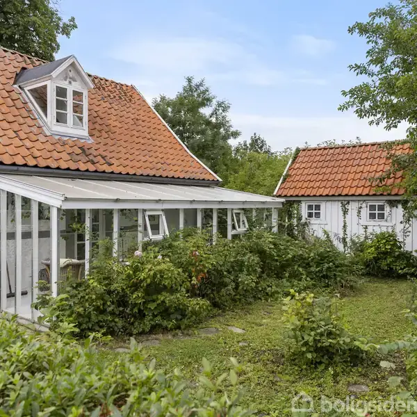 Et hvidt hus med rødt tegltag står i en frodig have. En udestue er ved siden af huset, omgivet af grønne buske og træer, under en klar blå himmel.