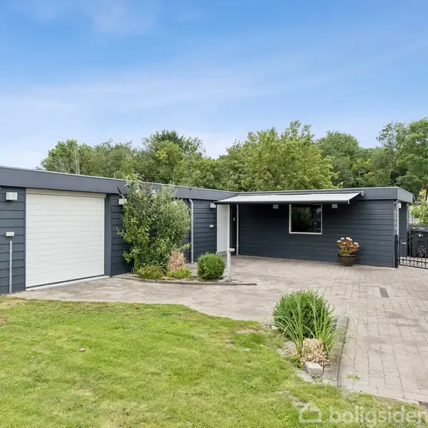 Et moderne, mørkegråt hus med garage ligger i en velplejet have med stenbelagt indkørsel. Huset er omgivet af grønne træer og en pæn græsplæne.