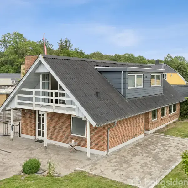 Et rødt murstenshus med sort skråtag har en balkon og store vinduer. Det står på en flisebelagt terrasse omgivet af græsplæne og andre huse i et grønt område.