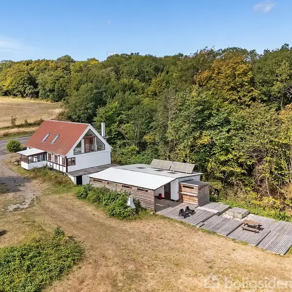 En hvid og rødmalet bolig med tilstødende udhus ligger ved en skovkant. Husets omgivelser inkluderer en have og en mark, mens et træterrasseområde er beliggende ved siden af bygningen.