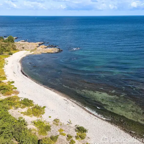 En sandstrand strækker sig langs kysten, omgivet af grøn vegetation under en blå himmel; havet fremstår roligt med svage bølger, mens klipper ses i baggrunden.