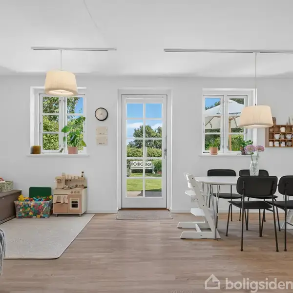 Stue med spisebord omgivet af sorte stole, belyst af moderne lamper. Legetøj og billeder ved siden af vindue og dør til have. Lyse vægge og trægulv skaber et lyst rum.