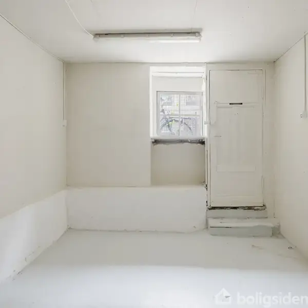 Et tomt, hvidt rum har en dør til højre ved et lille trin og et vindue over døren. Lyset er fra en loftmonteret lampe.