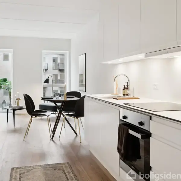 Køkkenet har en moderne ovn og kogeplade integreret i en hvid skabsvæg. I baggrunden står et rundt spisebord med sorte stole. Lyset er lyst og indbydende.