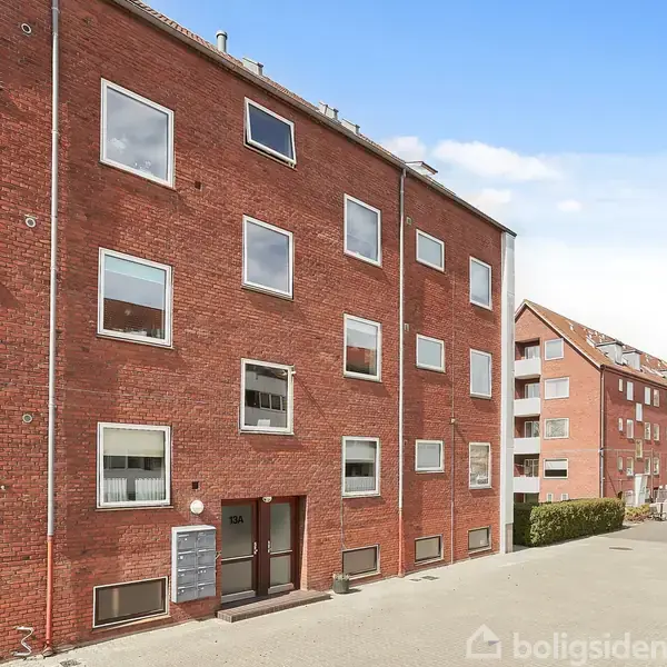 Rødt murstensbyggeri med flere vinduer, står stille i et åbent boligområde med flere lignende bygninger i baggrunden, eftermiddagssol skaber skygge på bygningens facade.