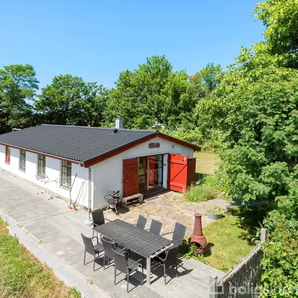 Hvidt hus med sorte tagsten og røde døre, der står ved flisebelagt terrasse med havemøbler. Omgivet af grønne træer i en solrig have.