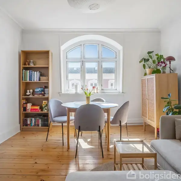 Et lyst værelse med et rundt spisebord og fire stole foran et stort buet vindue. En bogreol og et skab med planter står omkring bordet i en hyggelig stue.