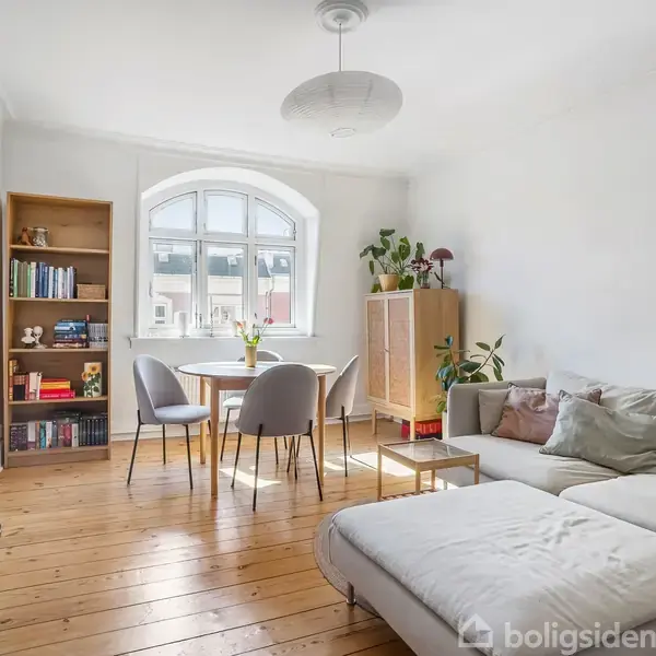 En stue med sofa og sofabord står ved siden af et stort vindue, der lader lys trænge ind. En spisebord med stole og en reol med bøger er til stede.