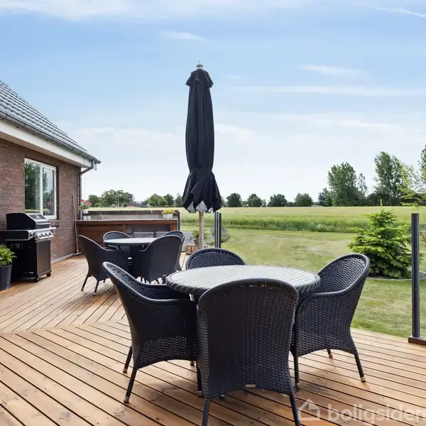 Terrassemøbler står på en trædækket terrasse med en grill i nærheden, foran et murstenshus. En parasol rejser sig over bordet. I baggrunden ses en stor, åben græsplæne.