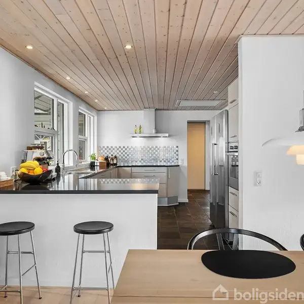Et moderne køkken med en sort bordplade og to høje skamler. Køkkenet har hvide skabe, ovn og indbygget komfur. På bordet er der en frugtskål. Træloft og lyse vægge skaber en lys atmosfære.