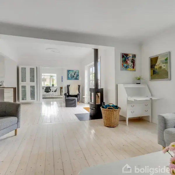 Lyst stueområde med grå stole og sofaer, pejs centralt placeret, skrivebord med billeder på væggen; træ- og plantede detaljer skaber en indbydende atmosfære.