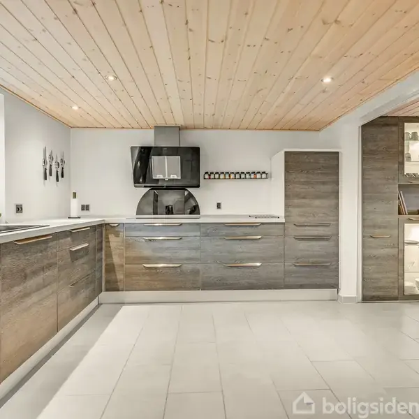 Et moderne køkken med mørke træskabe og lyse flisegulve. Skabe rummer en ovn og emhætte. Store vinduer giver naturligt lys med udsigt til grønne træer.