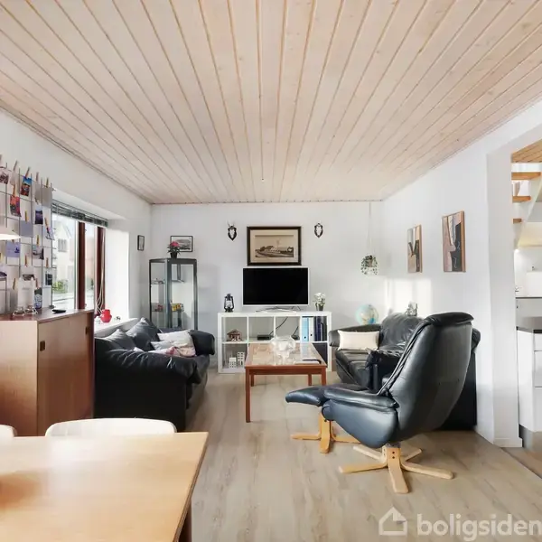 En stue med sofaer og lænestol, hvor en fladskærm står på en hylde mod bagvæggen. Rummet er lyst med store vinduer og træloft. Spisebord står i forgrunden.