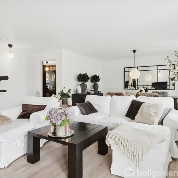 Hvid sofasæt danner en hjørnesektion, pyntet med tæpper og puder, omkring et sort sofabord med blomster og lysestage. Rummet har trægulve og moderne, minimalistisk indretning.