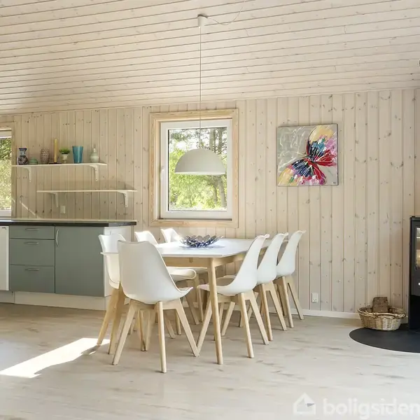 Et moderne køkken med lyse trævægge indeholder et spisebord med seks hvide stole. En brændeovn står i hjørnet, mens store vinduer giver udsigt til grønne omgivelser.