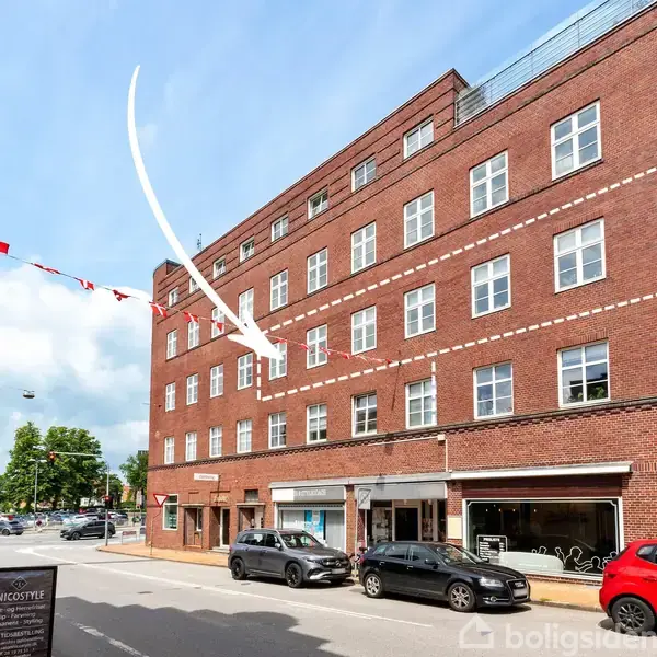 En rød murstensbygning med flere vinduer, hvor en pil peger op til en etage. Gaden er fyldt med parkerede biler og der hænger flag i snor over vejen.