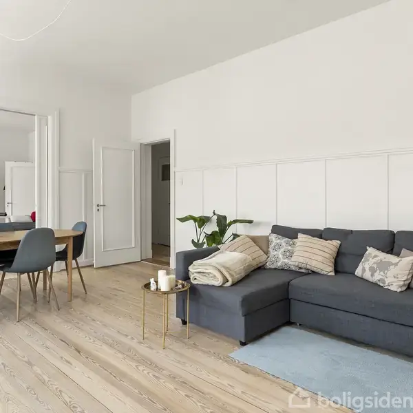Mørk sofa med puder placeret i moderne stue; rummet har lyse trægulve, spisebord med blå stole, og et stort spejl.