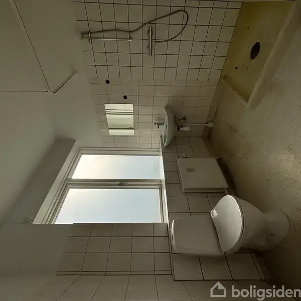 Toilet i funktionelt badeværelse med hvid flisevæg. Bruseområde til højre med bruser og armaturer. Stort vindue ved siden af håndvask og spejl. Lys og enkel indretning.