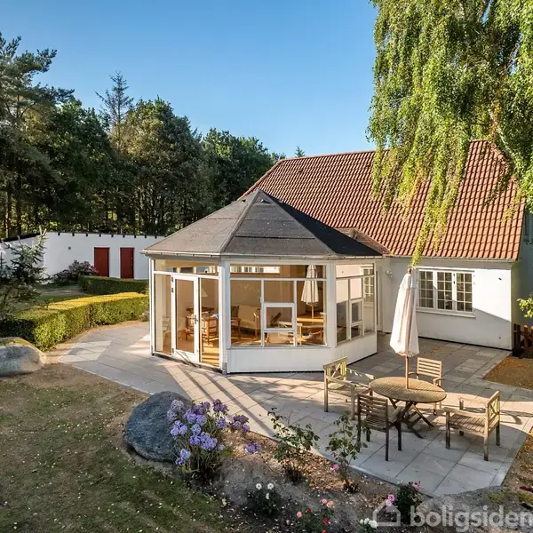 Hus med rødt tag og have. En rund, møbleret terrasse med parasol står ved siden af en glaspavillon. Skov omgiver ejendommen, der er fredelig og solrig.