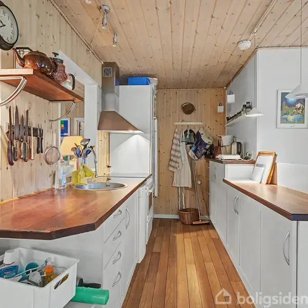 Køkkenbord med træoverflade er centralt i et hyggeligt køkkenmiljø. Knive hænger på væggen over bordet, omgivet af hvide skabe og trævægge med indbydende lys.