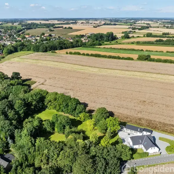 Hus ligger i åben landområde omgivet af marker og skove. Andre huse og en by ses i det fjerne, beliggende i et landligt landskab under en blå himmel.