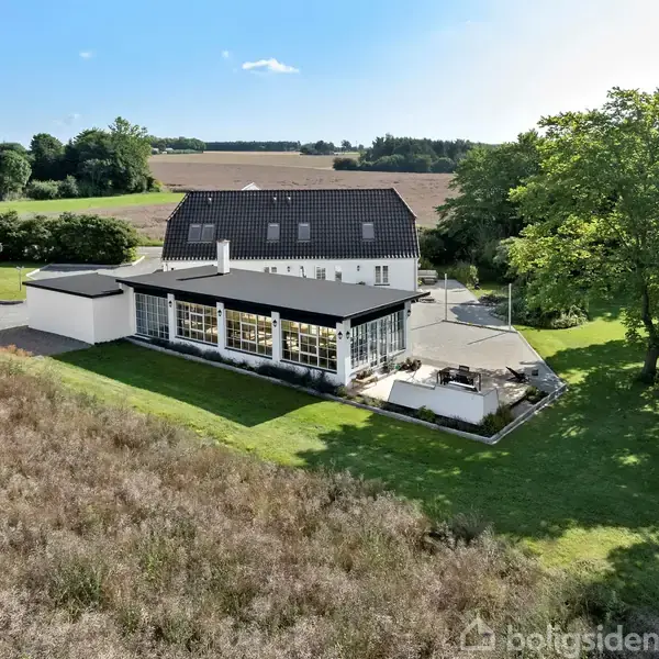 En stor villa med sort tegltag og hvid facade ligger omgivet af græsplæner og træer. En udestue strækker sig ud fra huset med udsigt til åbne marker.