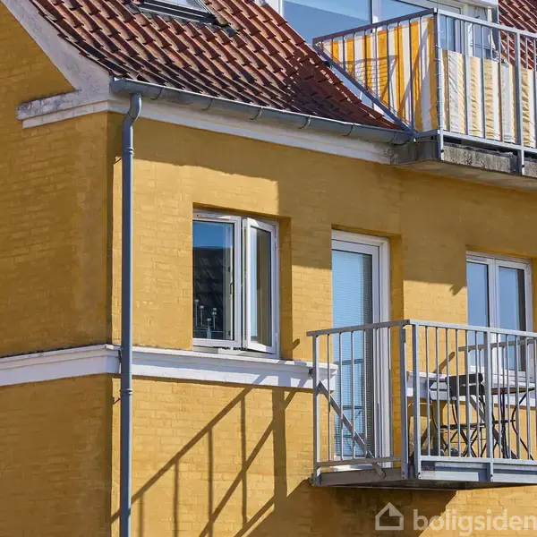 Gul murstensbygning med rødt tegltag, to altaner med metalgelændere. Soloplyst facade med tre hvide vinduer på øverste etage; urban boligmiljø.
