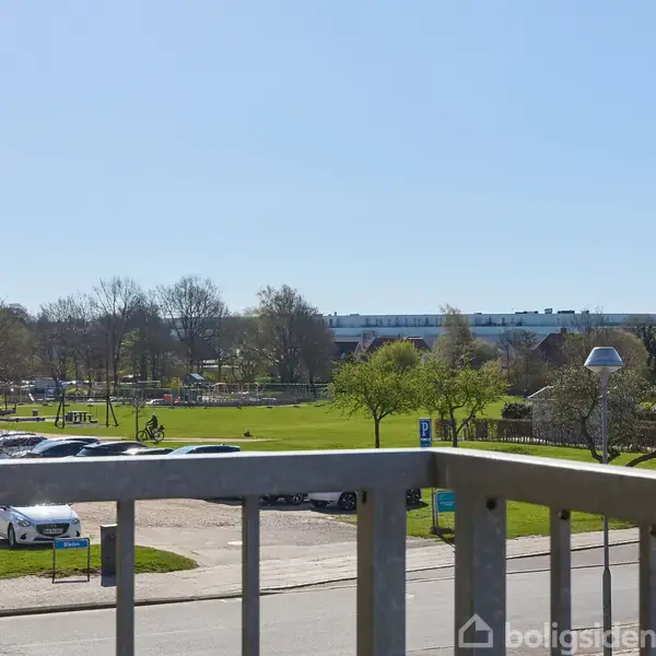 Udsigt fra altan med metalgelænder. Græsplæne og træer i parkområdet hvor folk cykler og går. Bygninger og biler i baggrunden under en klar blå himmel.