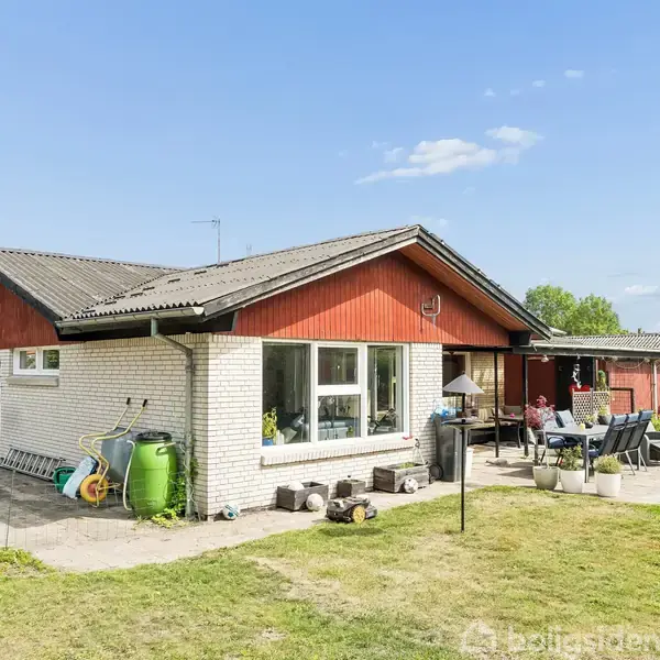 Huset er enfamilieshus med hvide mursten og rødt træværk. En terrasse med borde og stole har personer siddende udenfor. Græsplæne og hække omgiver huset.