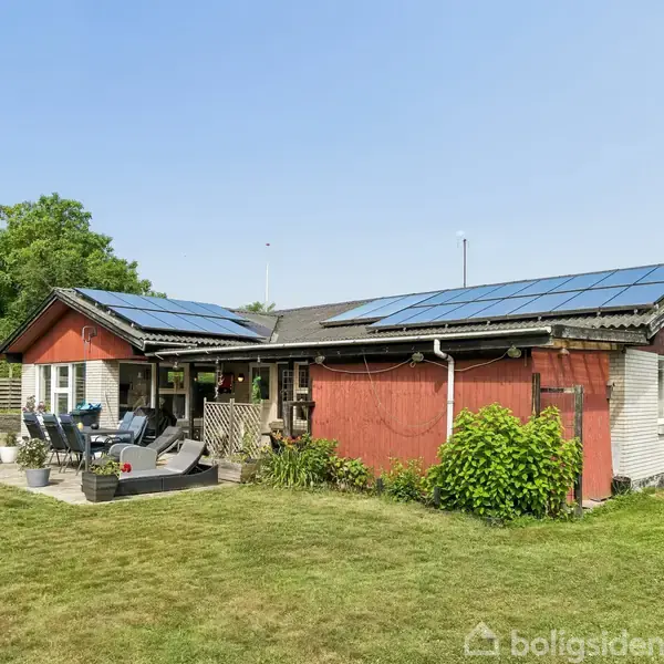 Et enfamiliehus med solpaneler på taget står i en grøn have. En terrasse med møbler er synlig foran huset, der er omgivet af træer og græsplæne.