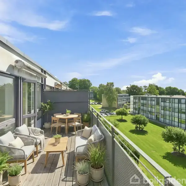 Balkon indrettet med hvidt loungesæt og planter. Det er en solrig dag, og balkonen har udsigt til grønne områder og moderne lejlighedsbygninger.