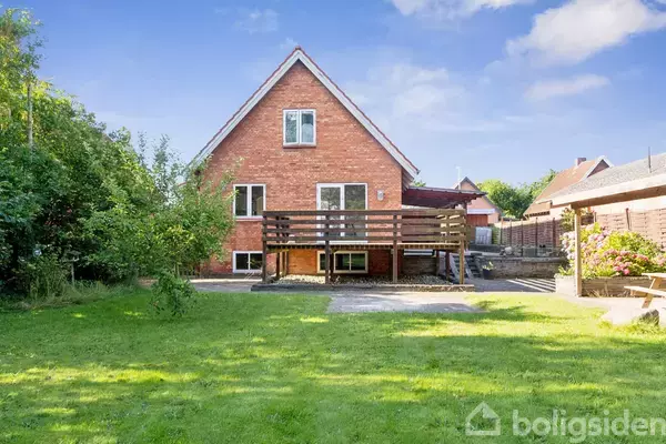 En rød murstensbygning med en stor træterrasse står i en have. Græsplæne og buske omgiver huset, der har en blå himmel i baggrunden.