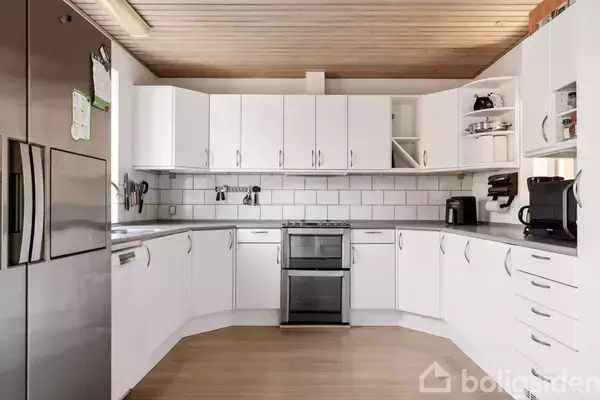 Et hvidt køkken med en L-formet bordplade. Der er hvide skabe, en fritstående ovn i midten og en sølvfarvet opvaskemaskine til venstre. Træloft og lysebrunt gulv.