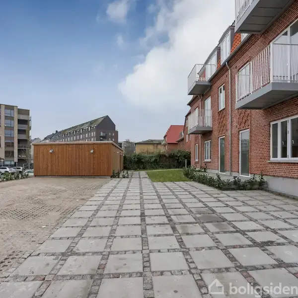 Rødt murstensbyggeri med altaner står stille langs en flisebelagt parkeringsplads. I baggrunden ses et træskur og lejlighedsbygninger under en klar blå himmel.