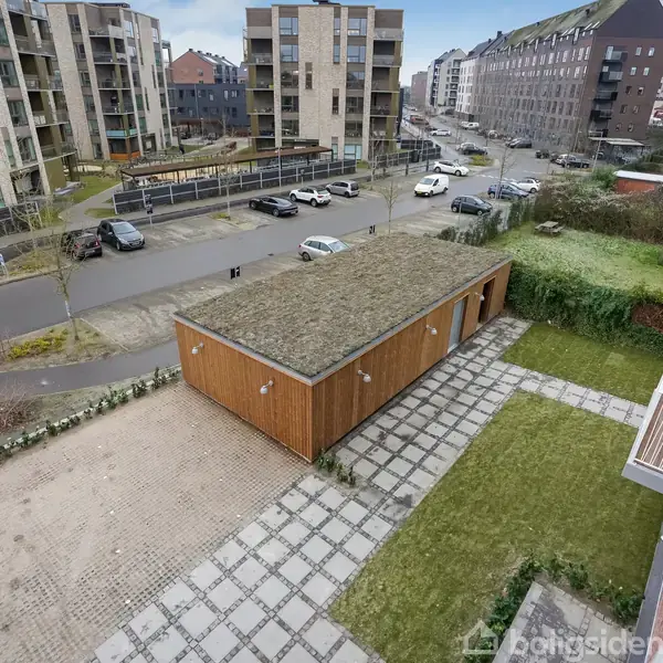 En græsbeklædt træbygning står stille i en gård omgivet af moderne lejlighedsbygninger i et byområde. Indkørsel og parkerede biler ses i nærheden.