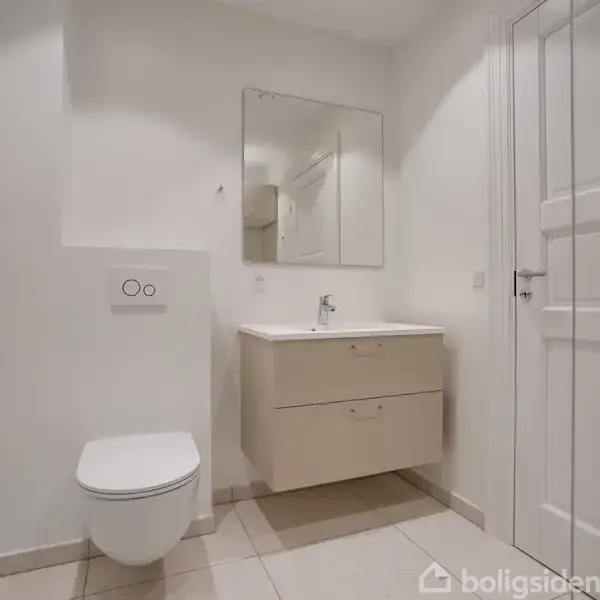 Toilet hænger på væggen; spejl og håndvask med skab under; hvidt, enkelt badeværelse. Der er lyse fliser på gulvet, og rummet er velbelyst.
