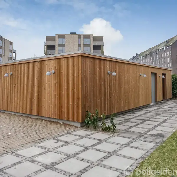 En rektangulær bygning med træbeklædning står stille ved siden af flisebelagt gangsti. Omgivet af moderne etagebygninger og græsareal i en boligkvarter.