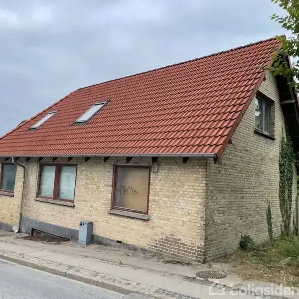 En gul murstensbygning står tom. Den har et rødt tegltag og et vindue med graffiti. Bygningen ligger ved en vej med græsset omgivelser.