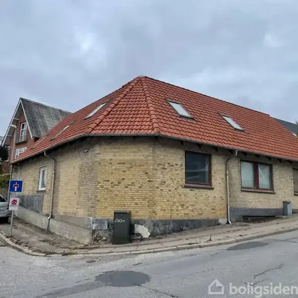 Et hjørnehus med gult murværk og rødt tegltag ligger ved en stille vej med kantsten. Der er tre tagvinduer på taget, og en parkeret bil ses i nærheden.