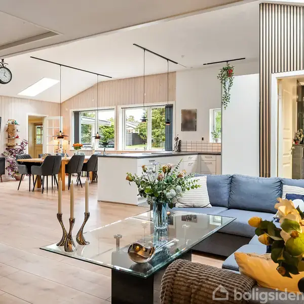 En moderne stue med en blå hjørnesofa står ved et glasbord med blomster og lysestager. I baggrunden er et åbent køkken med spisebord, stole og vinduer ud mod haven. Vægge med vinholdere ses til venstre.