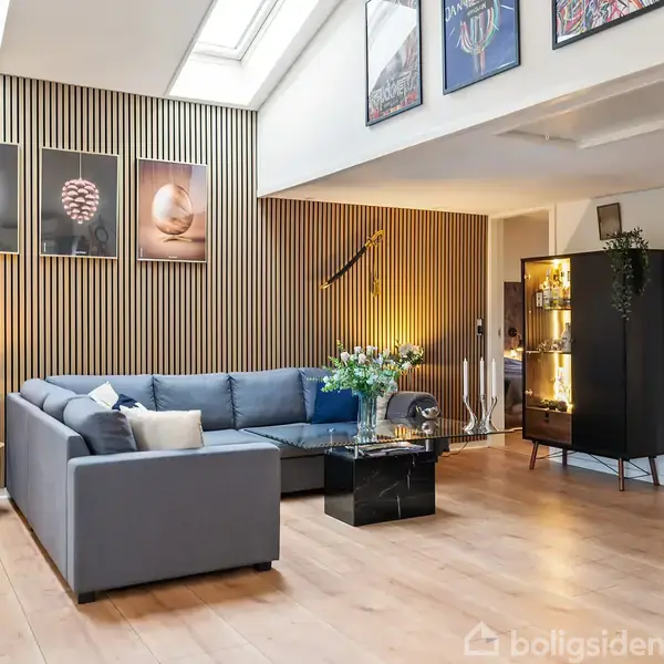 En moderne stue med en grå hjørnesofa, pyntet med puder. Der er kunst på væggen, plankegulv, og et sofabord med blomster i en vase. Ovenlys og planteudsmykning skaber lys og atmosfære.