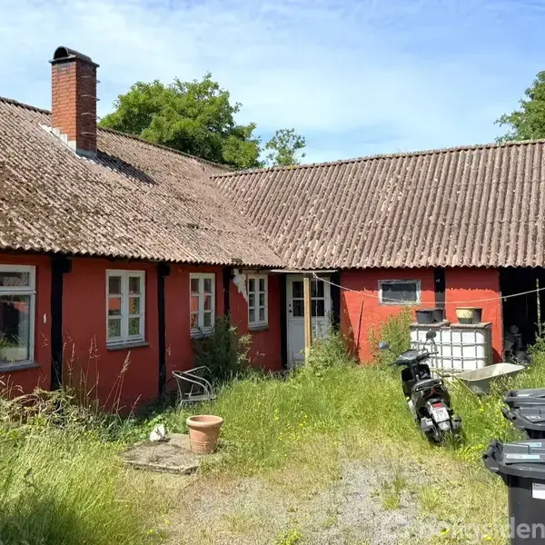 Rødt hus med skråt tag og skorsten. Foran står en scooter og nogle skraldespande i en gårdsplads med højt græs. Omkring ses grønne træer under en blå himmel.