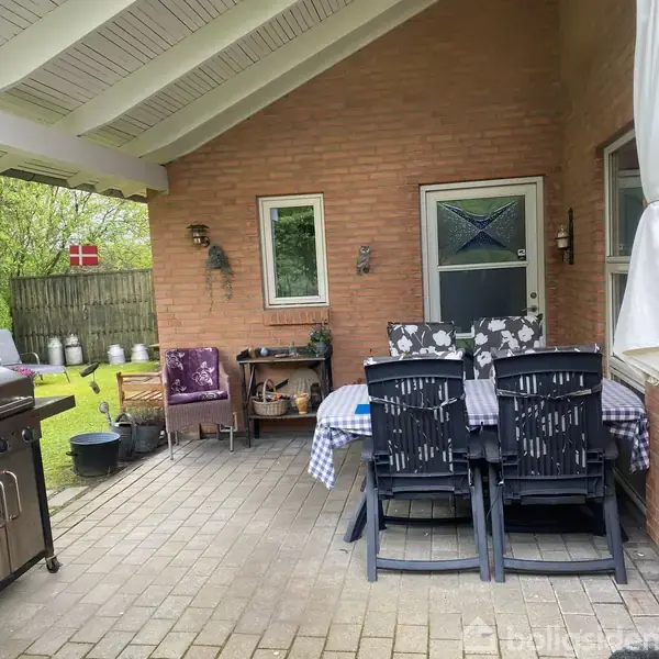 Terrasse med spisebord og stole under halvtag, omgivet af murstensvægge. Gasgrill står ved siden af, og danske flag kan ses i haven bagved.