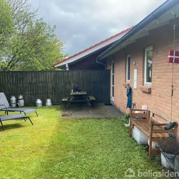 En baggård med græsplæne rummer to liggestole og et træbord med bænke. En rød murstensvæg med et lille dannebrogsflag hænger tæt på.