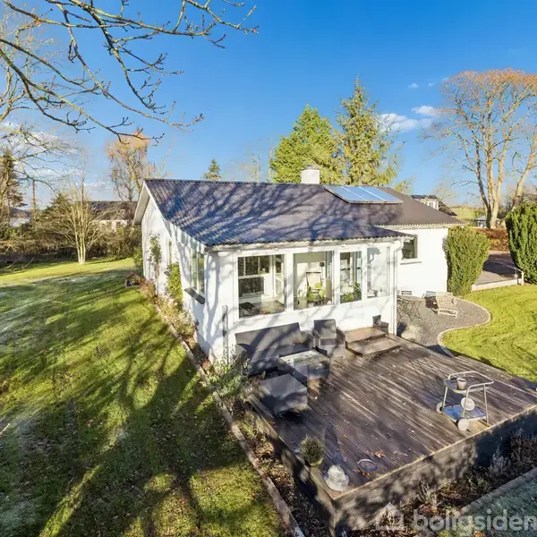 Hus står stille i sollys, omgivet af en stor have med græsplæne og træer. Træterrasse foran huset, himlen blå med enkelte skyer.