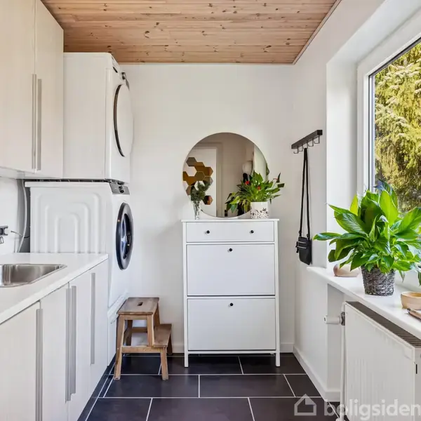 Vaskerum med vaskemaskine og tørretumbler stablet ved siden af vask. Et spejl og planter står på en hvid kommode. Store vinduer viser træer udenfor.
