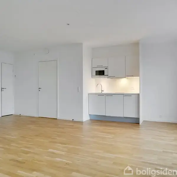 Et lyst rum med trægulv, hvor et lille køkken med hvide skabe og indbygget mikrobølgeovn er monteret på væggen. To døre fører til andre rum, og der er store vinduer.