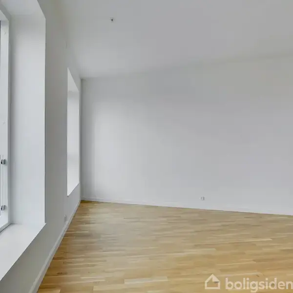 Tomt rum med trægulv, store vinduer til venstre giver naturligt lys, omgivet af hvide vægge, skaber en lys, minimalistisk atmosfære.