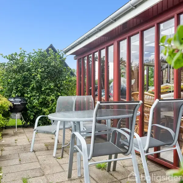 Et rundt havebord med fire stole står på en flisebelagt terrasse. En grill er placeret til venstre, omgivet af grønne planter foran et glashus med røde rammer.
