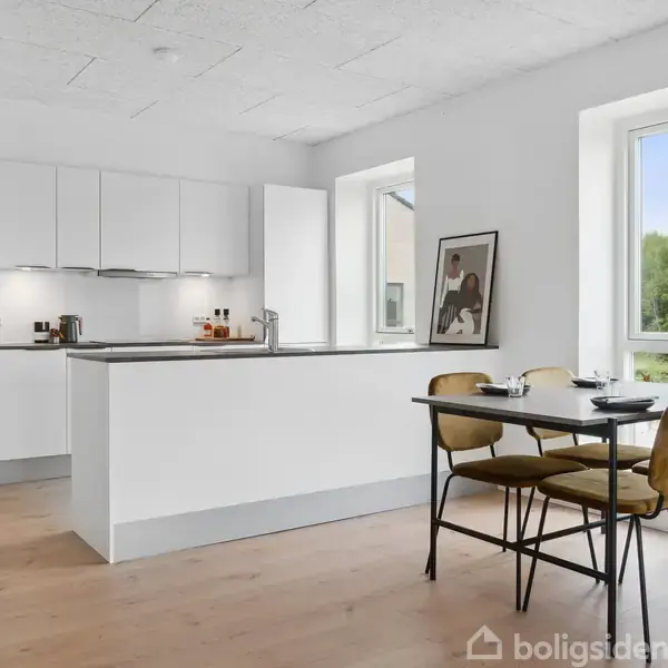 Et moderne køkken med hvide skabe og en køkkenø. Et spisebord med fire stole står ved et vindue, der giver udsigt til grønne omgivelser. En plakat er lænet op ad væggen.
